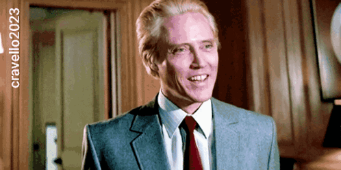 A Man In A Suit And Tie Is Smiling In Front Of A Sign That Says Cravello2023 GIF