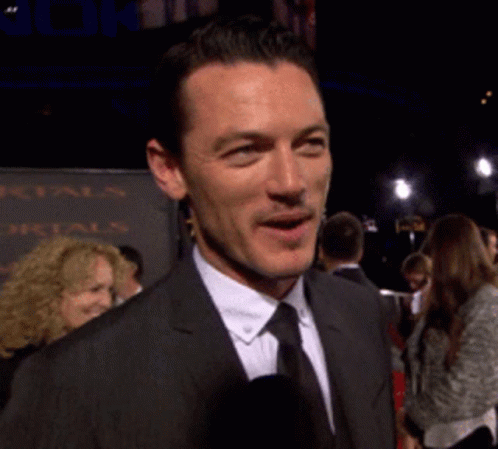 A Man In A Suit And Tie Is Smiling In Front Of A Sign That Says Mortals GIF