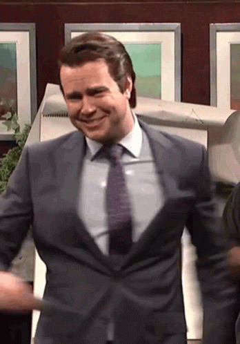 A Man In A Suit And Tie Is Smiling In Front Of A Wall Of Pictures GIF