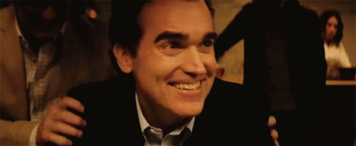 A Man In A Suit And Tie Is Smiling While Being Hugged By Another Man In A Dark Room . GIF
