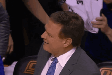 A Man In A Suit And Tie Is Smiling While Sitting In A Stadium GIF