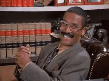 A Man In A Suit And Tie Is Smiling While Sitting In Front Of A Bookshelf . GIF