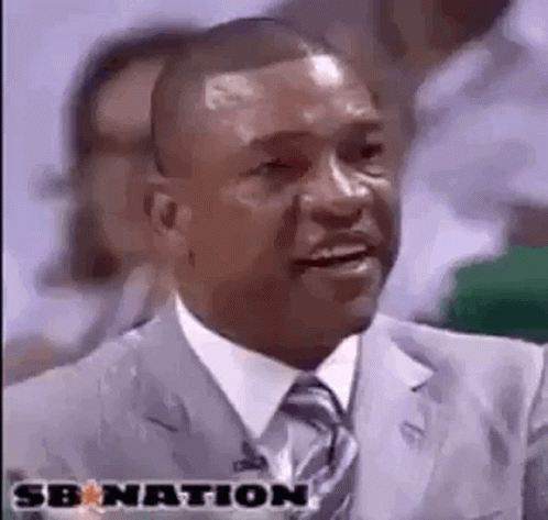 A Man In A Suit And Tie Is Smiling While Sitting In Front Of A Crowd Of People . GIF