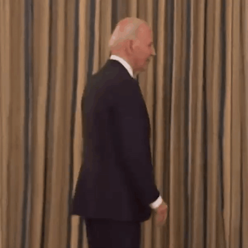 A Man In A Suit And Tie Is Smiling While Standing In Front Of A Striped Curtain . GIF