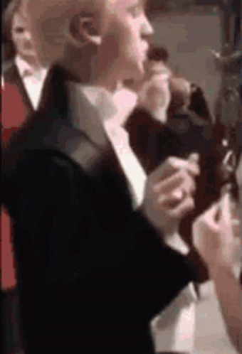 A Man In A Suit And Tie Is Smoking A Cigarette While Standing In A Crowd Of People . GIF