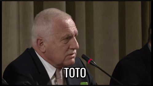 A Man In A Suit And Tie Is Speaking Into A Microphone And The Word Toto Is On The Screen Behind Him GIF