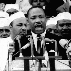 A Man In A Suit And Tie Is Speaking Into A Microphone In Front Of A Crowd Of People . GIF