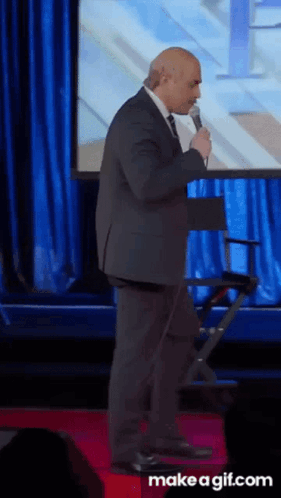 A Man In A Suit And Tie Is Speaking Into A Microphone On A Red Carpet GIF