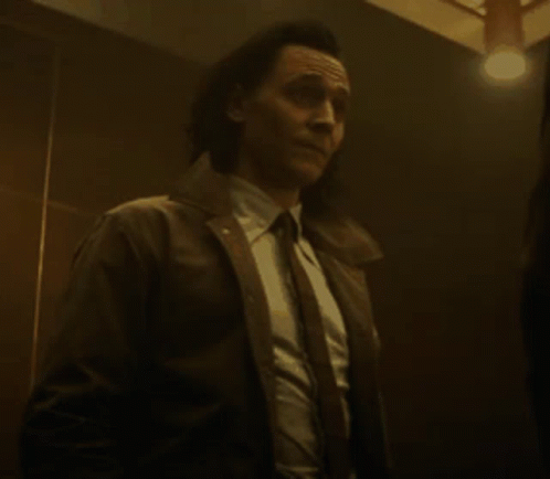 A Man In A Suit And Tie Is Standing In A Dark Room With A Light On . GIF