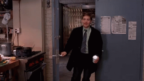 A Man In A Suit And Tie Is Standing In A Kitchen Holding A Cup And A Flashlight . GIF