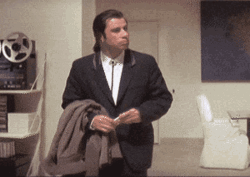 A Man In A Suit And Tie Is Standing In A Room Holding His Jacket GIF