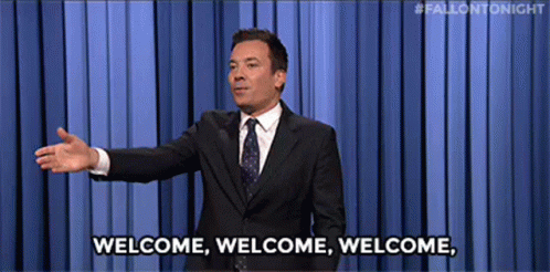 A Man In A Suit And Tie Is Standing In Front Of A Blue Curtain And Says Welcome . GIF