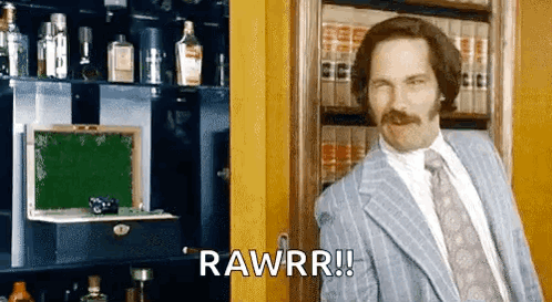 A Man In A Suit And Tie Is Standing In Front Of A Bookshelf And Saying Rawrr ! GIF