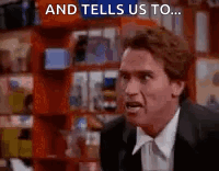 A Man In A Suit And Tie Is Standing In Front Of A Bookshelf And Talking To Someone . GIF
