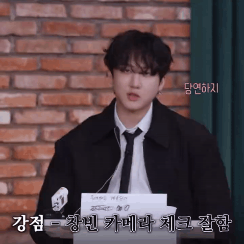 A Man In A Suit And Tie Is Standing In Front Of A Brick Wall And Holding A Piece Of Paper With Korean Writing On It GIF