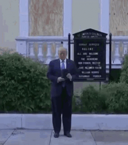 A Man In A Suit And Tie Is Standing In Front Of A Building With A Sign . GIF