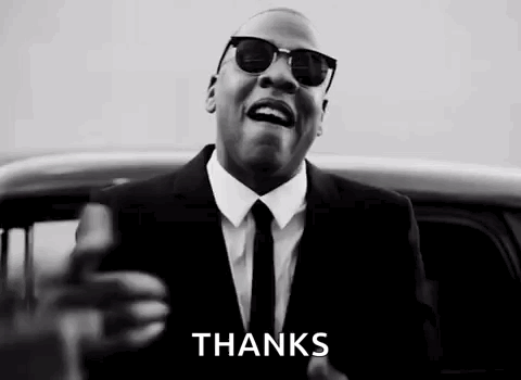 A Man In A Suit And Tie Is Standing In Front Of A Car And Giving A Thanks Sign . GIF