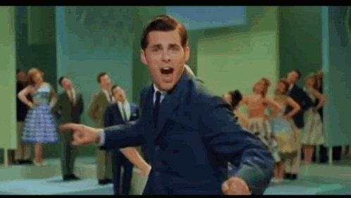 A Man In A Suit And Tie Is Standing In Front Of A Crowd Of People . GIF