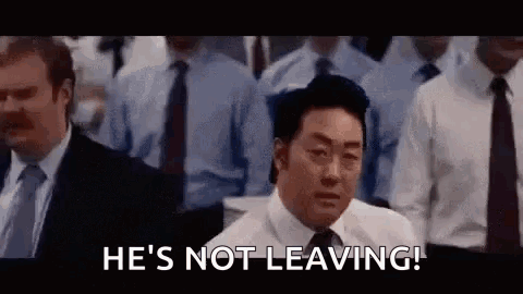 A Man In A Suit And Tie Is Standing In Front Of A Crowd Of People And Says `` He 'S Not Leaving ! '' GIF