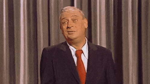 A Man In A Suit And Tie Is Standing In Front Of A Curtain And Making A Funny Face . GIF
