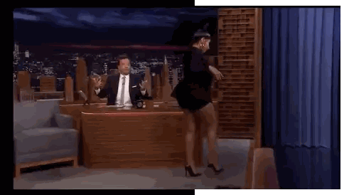 a man in a suit and tie is standing in front of a desk while a woman is dancing in front of a curtain