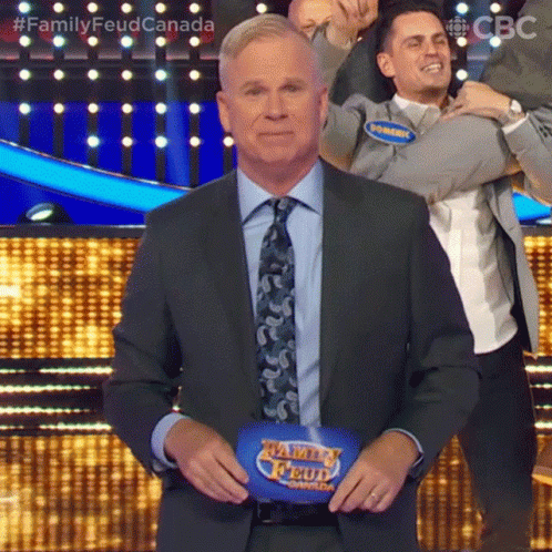 A Man In A Suit And Tie Is Standing In Front Of A Family Feud Podium . GIF