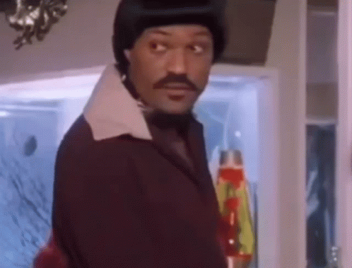 A Man In A Suit And Tie Is Standing In Front Of A Lava Lamp And Looking At The Camera . GIF
