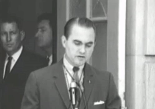 A Man In A Suit And Tie Is Standing In Front Of A Microphone GIF