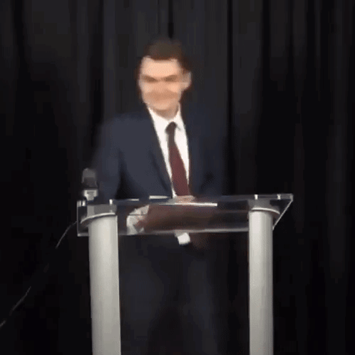 A Man In A Suit And Tie Is Standing In Front Of A Podium With His Arms Outstretched . GIF