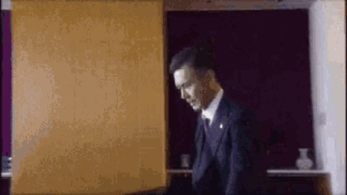 A Man In A Suit And Tie Is Standing In Front Of A Purple Wall In A Room . GIF