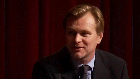A Man In A Suit And Tie Is Standing In Front Of A Red Curtain And Smiling . GIF