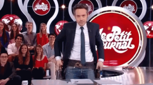 A Man In A Suit And Tie Is Standing In Front Of A Red Sign That Says Le Petit Journal GIF