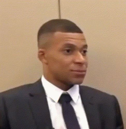 A Man In A Suit And Tie Is Standing In Front Of A Wall And Looking At The Camera . GIF