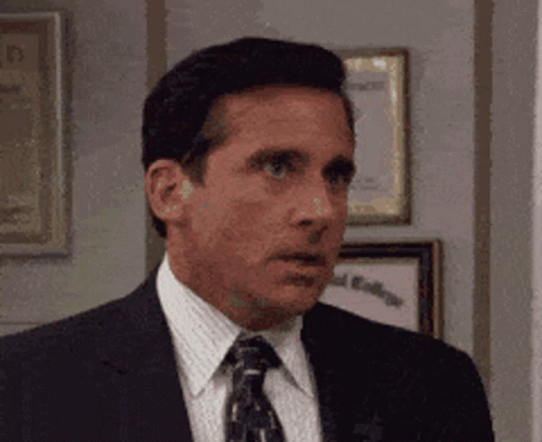 A Man In A Suit And Tie Is Standing In Front Of A Wall With Framed Pictures On It . GIF