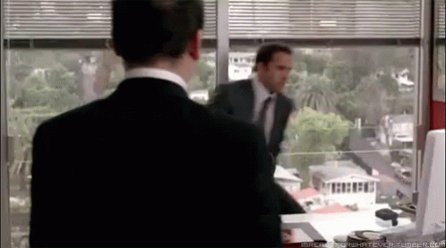 A Man In A Suit And Tie Is Standing In Front Of A Window Looking Out GIF