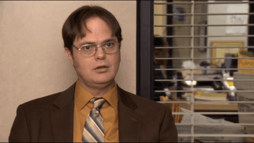 A Man In A Suit And Tie Is Standing In Front Of A Window With Blinds GIF