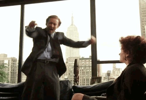 A Man In A Suit And Tie Is Standing In Front Of A Woman In A Chair GIF