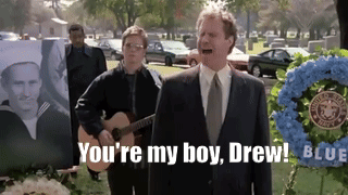 A Man In A Suit And Tie Is Standing In Front Of A Wreath That Says ' You 'Re My Boy Drew ' GIF