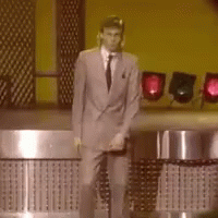 A Man In A Suit And Tie Is Standing In Front Of A Yellow Wall GIF