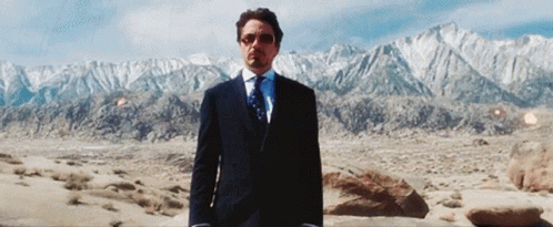 A Man In A Suit And Tie Is Standing In The Desert With Mountains In The Background GIF