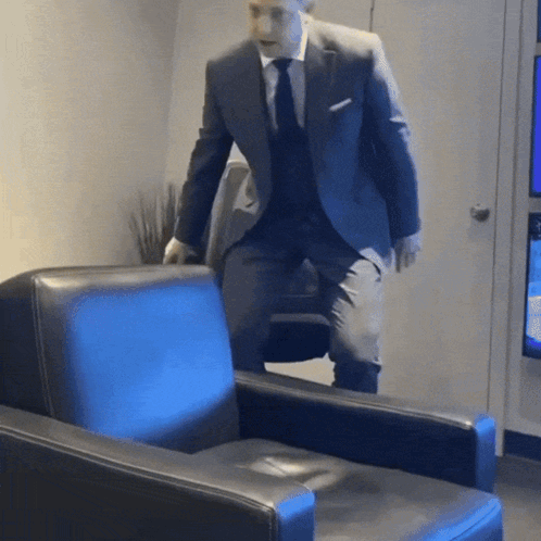 A Man In A Suit And Tie Is Standing Next To A Blue Chair GIF