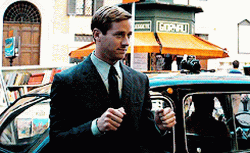 A Man In A Suit And Tie Is Standing Next To A Car In Front Of A Store That Says Gornelli GIF