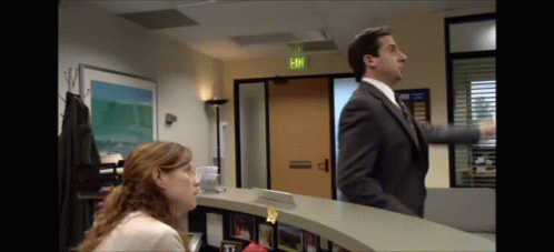 A Man In A Suit And Tie Is Standing Next To A Woman At A Reception Desk In Front Of An Exit Sign GIF