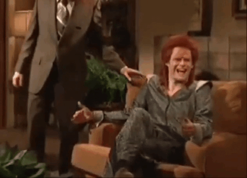 A Man In A Suit And Tie Is Standing Next To A Woman In A Chair . GIF