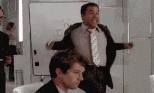 A Man In A Suit And Tie Is Standing Next To Another Man In A Chair . GIF