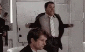 A Man In A Suit And Tie Is Standing Next To Another Man In A Chair . GIF