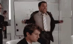 A Man In A Suit And Tie Is Standing Next To Another Man In A Chair In Front Of A Whiteboard . GIF