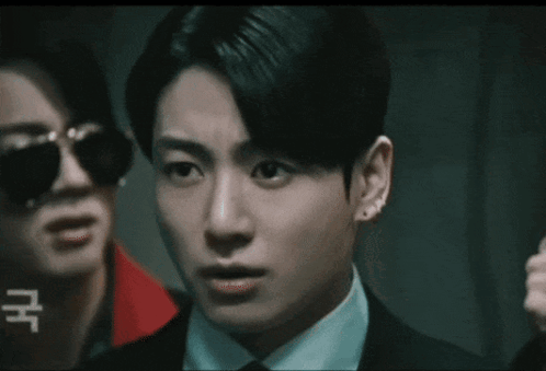 A Man In A Suit And Tie Is Standing Next To Another Man Wearing Sunglasses And Earrings . GIF
