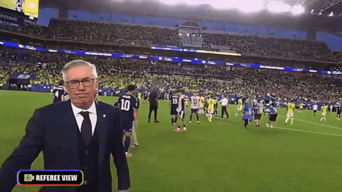 A Man In A Suit And Tie Is Standing On A Soccer Field With A Referee View Button On The Bottom GIF