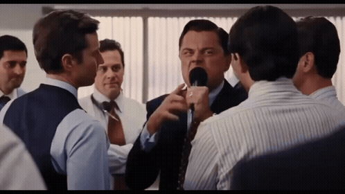 A Man In A Suit And Tie Is Talking Into A Microphone While Another Man Looks On GIF